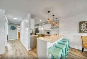A modern kitchen with white cabinetry, a central island with bar stools, pendant lighting, and a view into the adjacent dining area. - Green Reef 6•Remodeled•Private Beach•Beach Service & Beach Gear•3 Patios•Games (Destin)