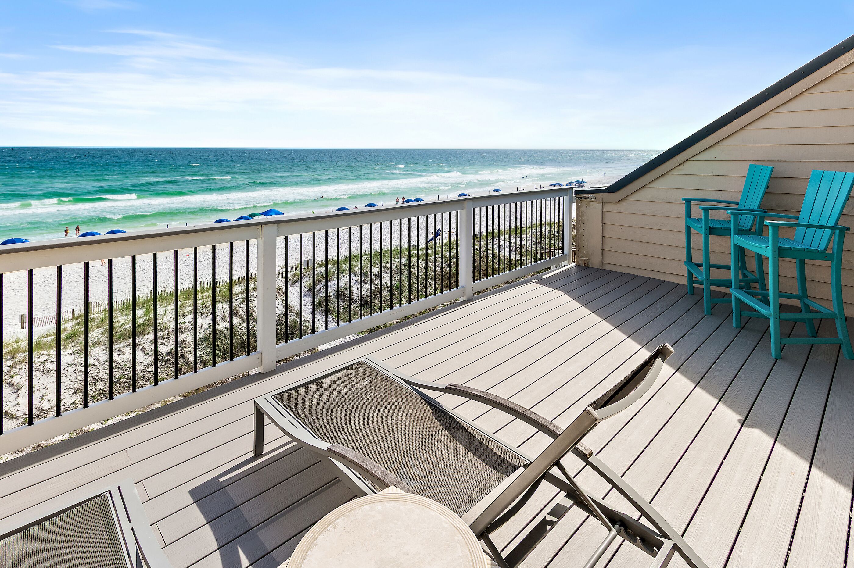 A wooden deck with a view of the beach and ocean, featuring a railing, a couple of chairs, and a table.