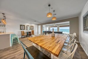 A spacious dining area with a wooden table and white chairs, a kitchen area with white cabinets, and a view of the ocean through large windows. - Green Reef 6•Remodeled•Private Beach•Beach Service & Beach Gear•3 Patios•Games (Destin)