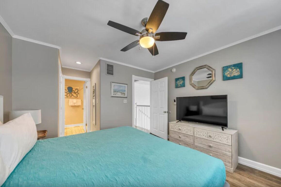 A bedroom with a bed, a ceiling fan, a TV on a dresser, and a door leading to another room.
