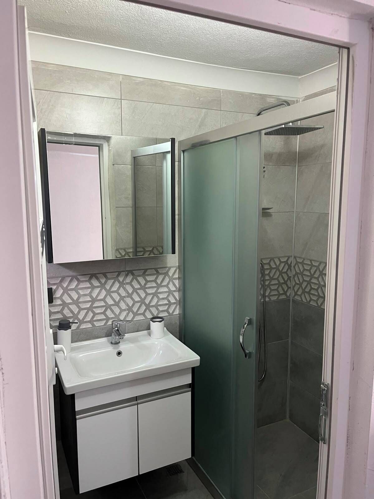 Une salle de bain avec un lavabo blanc, un miroir, un espace douche et un mur recouvert de carreaux décoratifs.