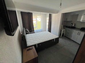 A compact room with a bed, a small desk, a kitchenette, and a window with curtains.