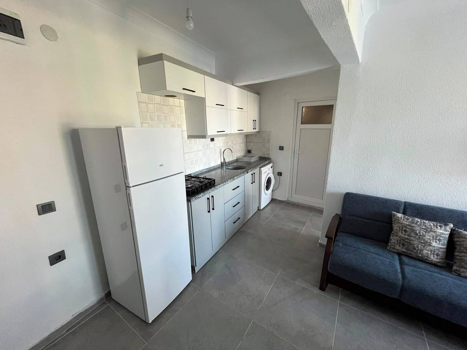 A compact kitchen with white cabinets, a built-in washing machine, and a blue sofa in the adjacent living area.