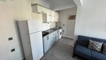A compact kitchen with white cabinets, a built-in washing machine, and a blue sofa in the adjacent living area.