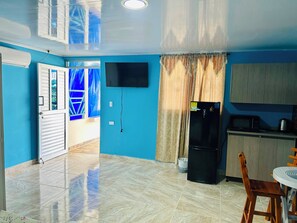 A room with a tiled floor, a black refrigerator, a wooden cabinet, a television mounted on the wall, and a door with a window.