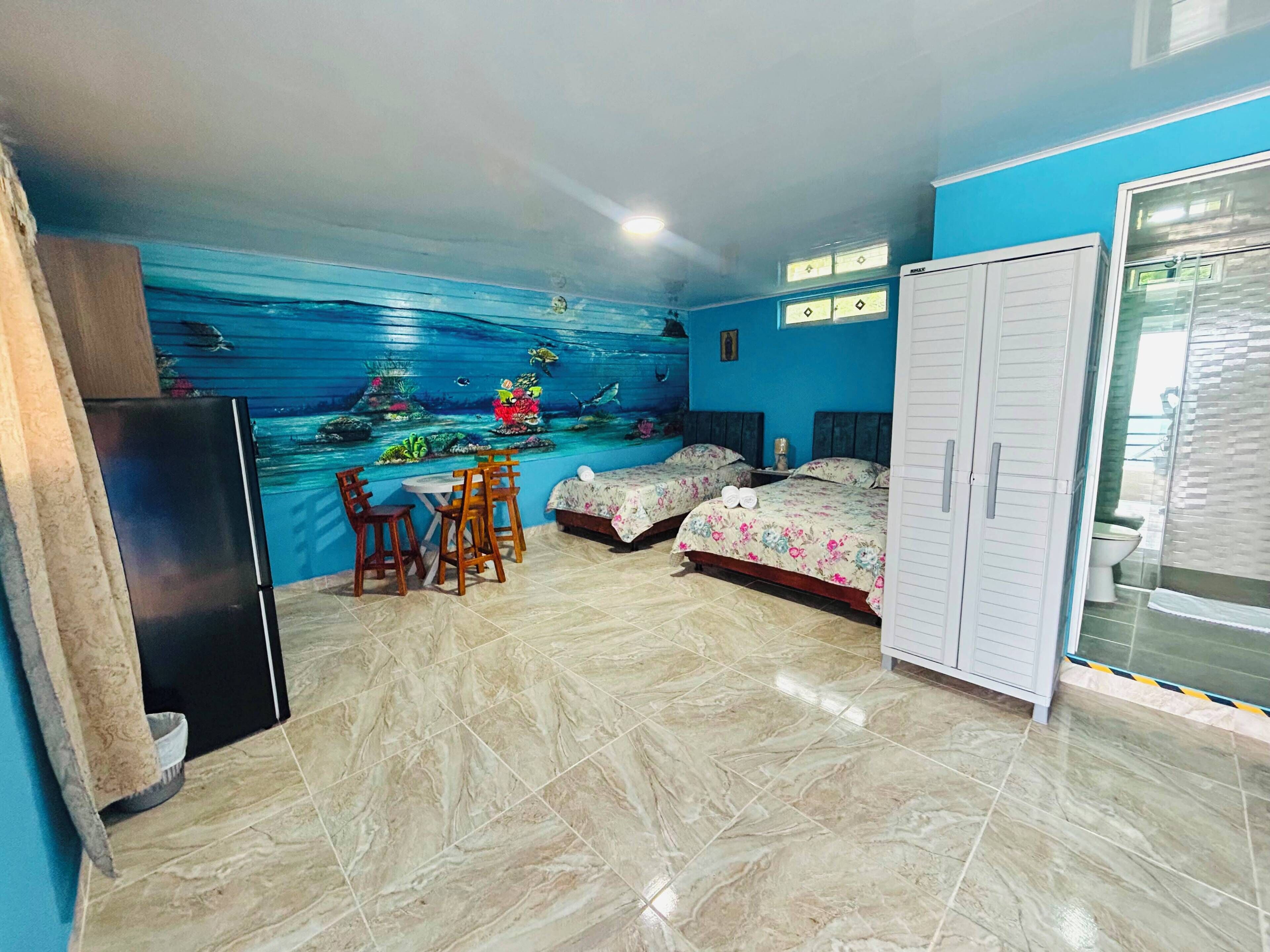 Habitación con un mural de una escena submarina, dos camas con ropa de cama floral, un armario blanco y un suelo de baldosas.