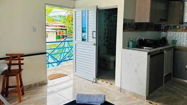 Cocina moderna con estufa integrada, un lavamanos y armarios. Se observa una zona de comedor con mesa y sillas. Se ve un baño a través de un umbral abierto.