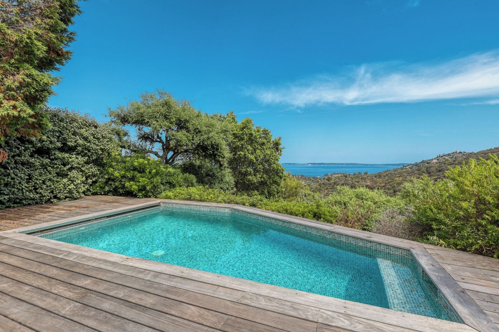 A rectangular swimming pool with clear blue water, surrounded by a wooden deck, set against a backdrop of lush greenery and a distant view of the sea.