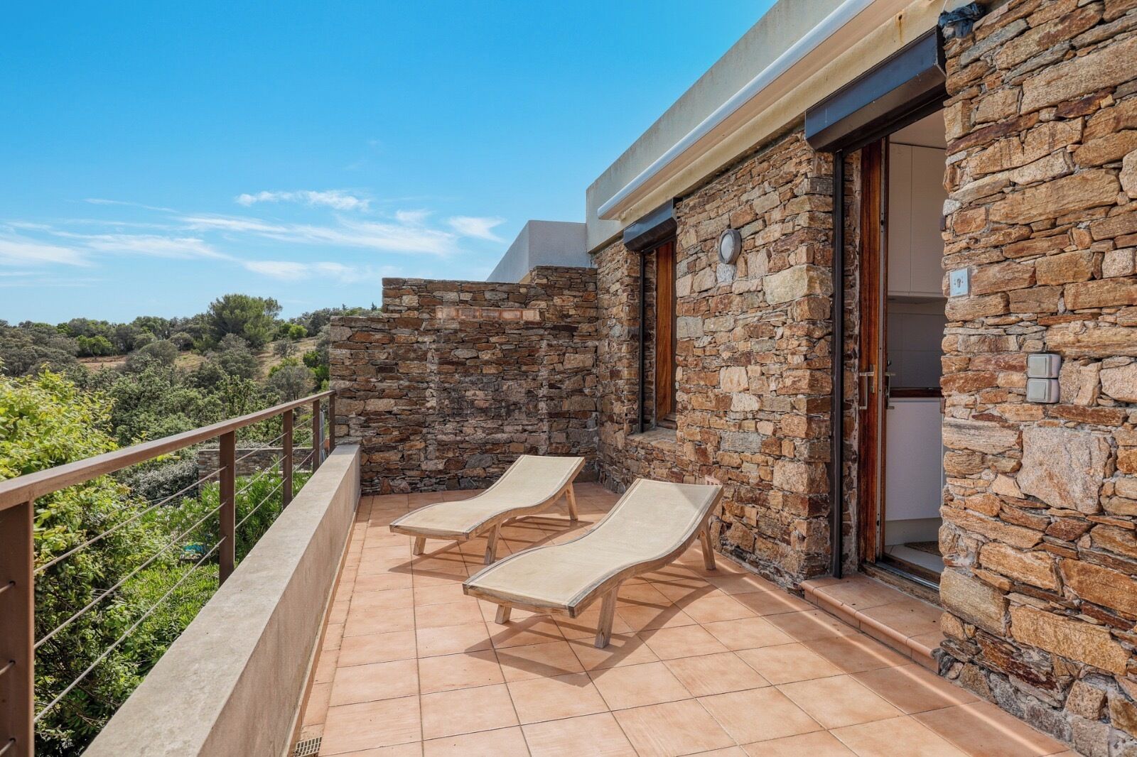 A balcony with stone walls, two lounge chairs, and a view of the countryside.