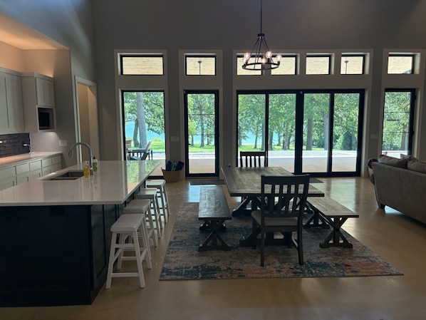 A modern kitchen with a central island, a dining area with a bench and chairs, and large windows offering views of trees and a body of water.