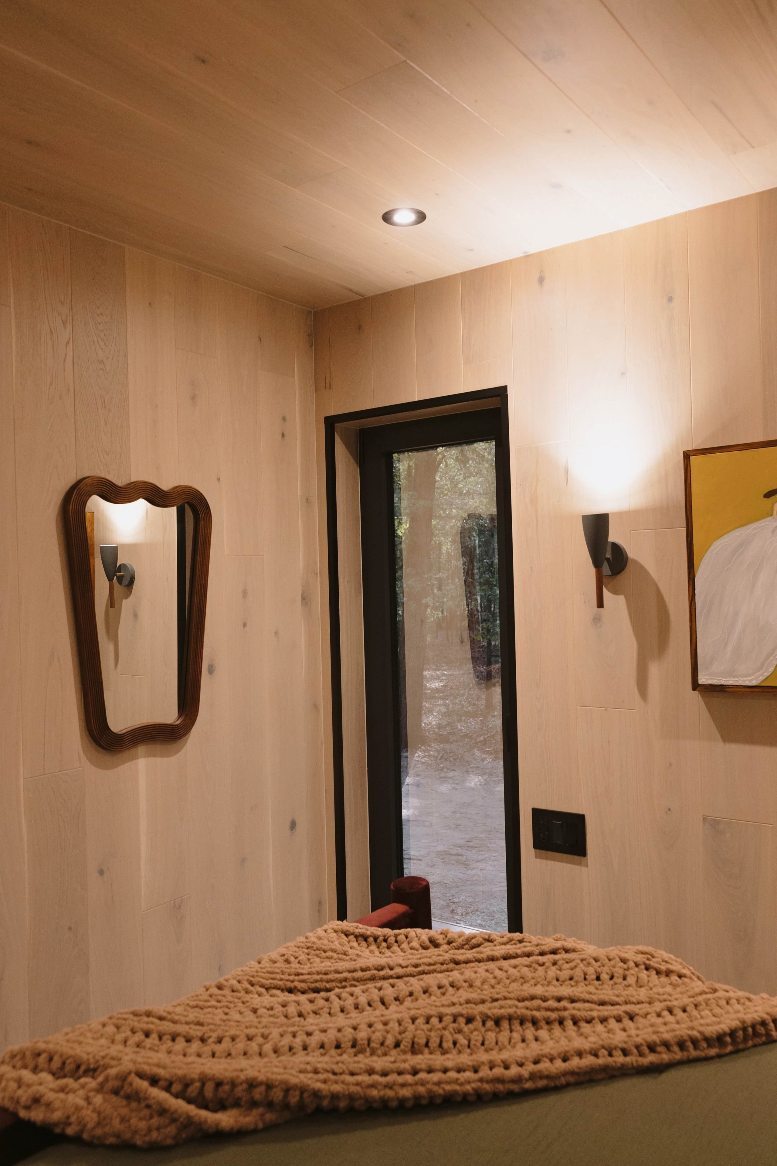 A room with wooden walls, a bed with a knitted blanket, a mirror, a door leading outside, and a wall-mounted light fixture.
