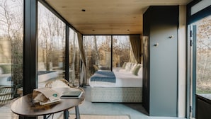 Ein modernes Interieur mit einem großen Fenster, einem Bett mit gemusterten Textilien, einem Tisch mit einer Tasche und einem Stuhl.