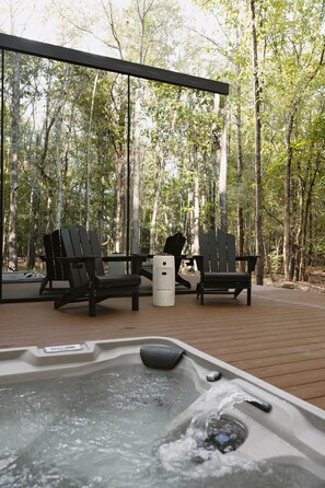 An outdoor hot tub area with two lounge chairs and a small table, surrounded by a wooden deck and trees. - Glass House IV tucked in the East Texas forest (Gregg County)