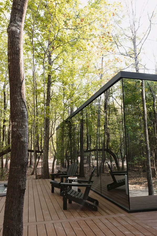 Una terraza de madera con sillas Adirondack y una estructura con paredes de cristal, rodeada de un bosque.