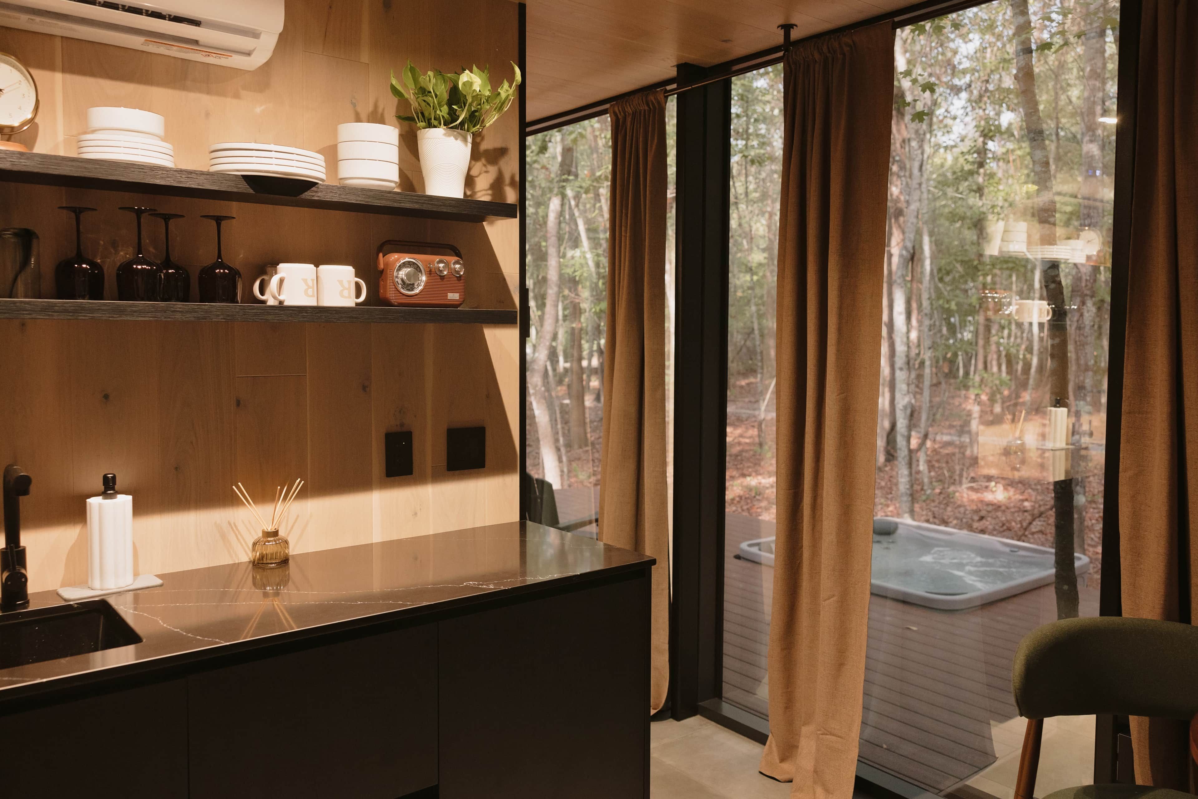Une cuisine moderne avec un plan de travail en bois, des étagères remplies de vaisselle, une plante en pot et une vue sur une forêt à travers de grandes fenêtres.