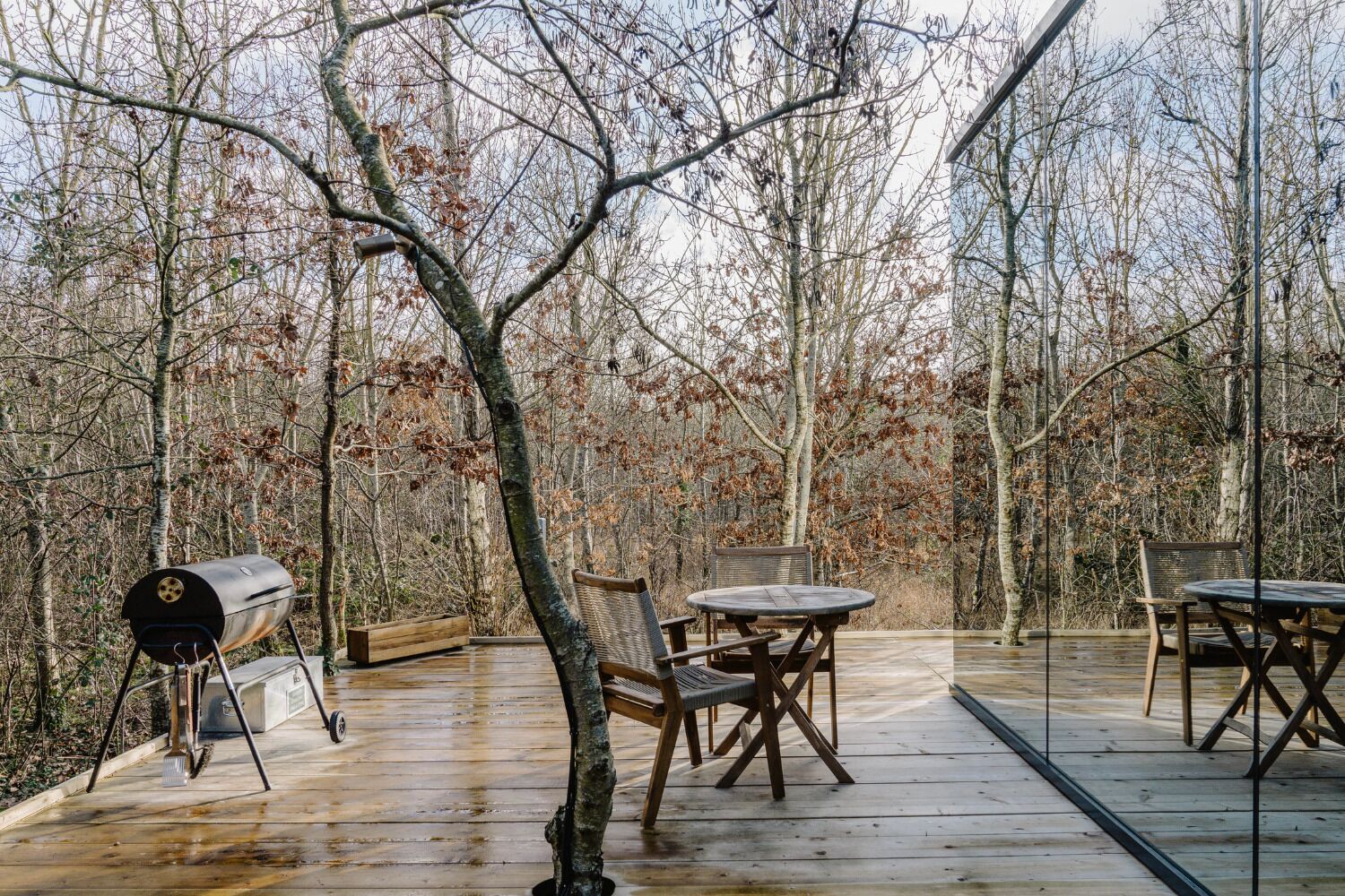 A wooden deck with a grill, tables, and chairs, surrounded by trees.