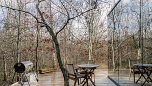 Eine Holzterrasse mit Grill, Tischen und Stühlen, umgeben von Bäumen.