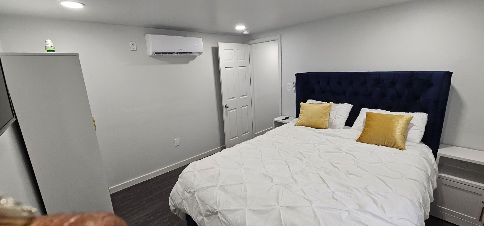 A bedroom with a bed, bedside tables, a wardrobe, a door, and an air conditioning unit.