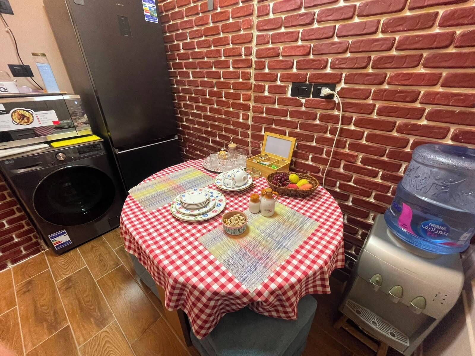 A compact kitchen with a brick wall, a washing machine, a water dispenser, and a table set with plates, cups, and a basket of fruit.