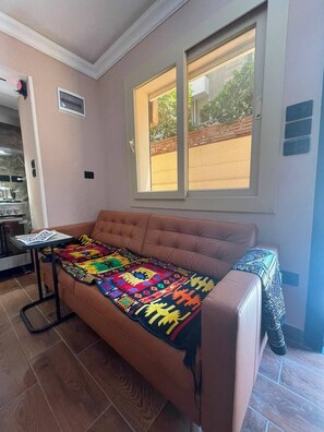 Ein Zimmer mit braunem Sofa, einer bunten Decke, einem Fenster mit Blick und einem kleinen Tisch mit einer Zeitschrift.