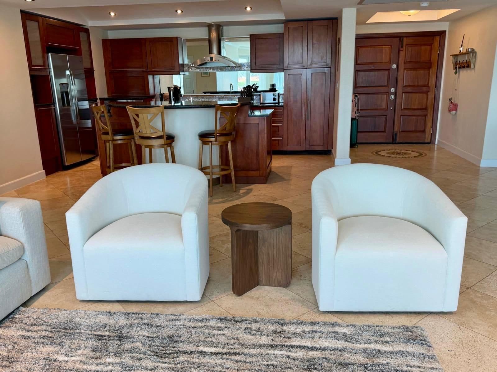 Una cocina moderna con armarios de madera, un refrigerador de acero inoxidable y una isla central. En la zona de estar hay dos sillones blancos y un taburete de madera.