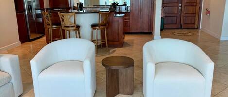 Una cocina moderna con armarios de madera, un refrigerador de acero inoxidable y una isla central. En la zona de estar hay dos sillones blancos y un taburete de madera.