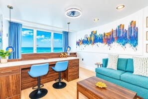 A modern living room with a cityscape mural, a sofa, and a view of the ocean through large windows. - Oceanfront Blue Haven at Yachtsman by Skywheel! (Myrtle Beach)