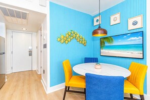 A room with a blue wall, a round table with yellow chairs, a ceiling fan, and a framed picture of a beach. - Oceanfront Blue Haven at Yachtsman by Skywheel! (Myrtle Beach)