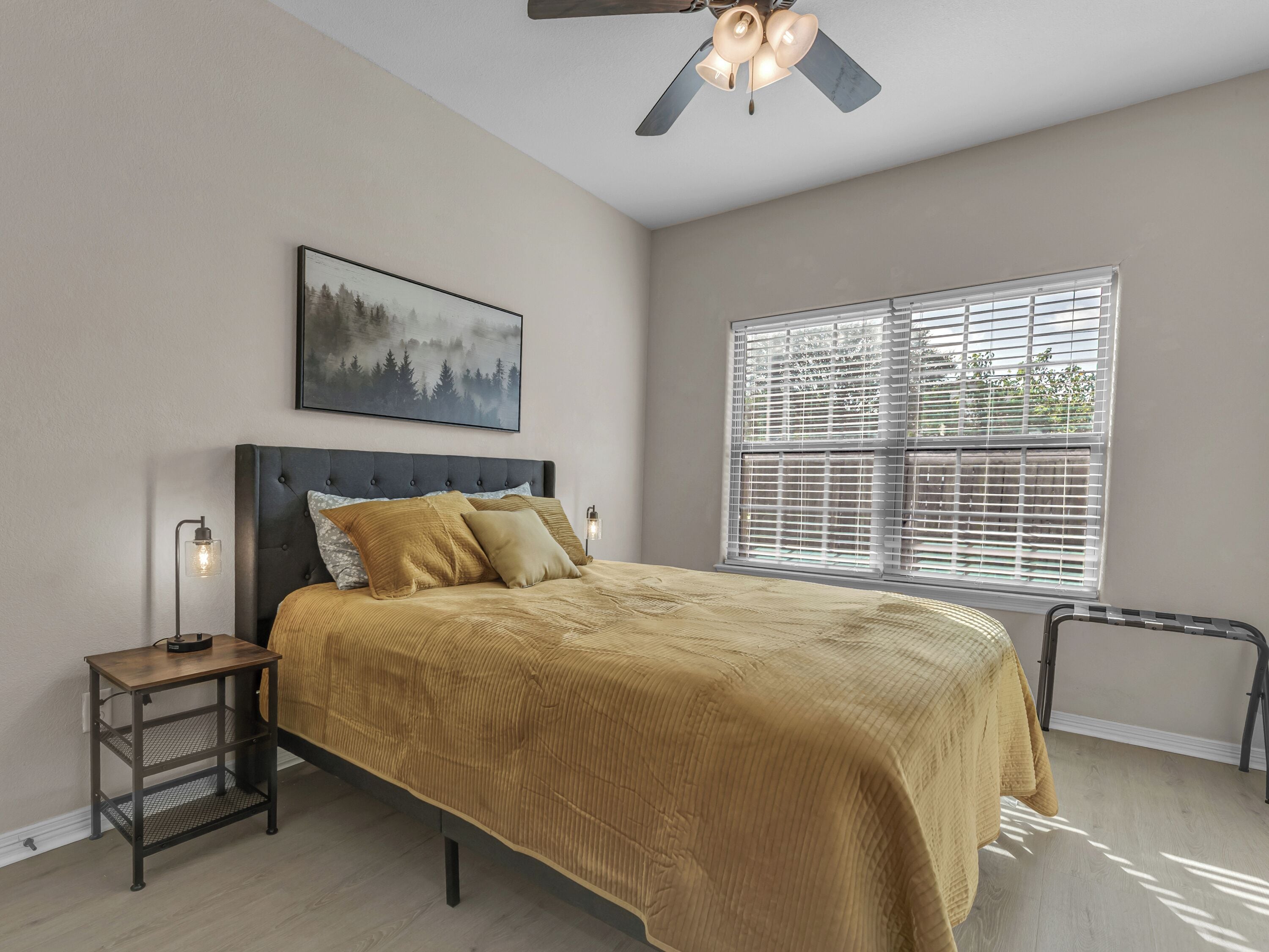 A bedroom with a bed, a nightstand, a wall-mounted picture, a ceiling fan, and a window with blinds.