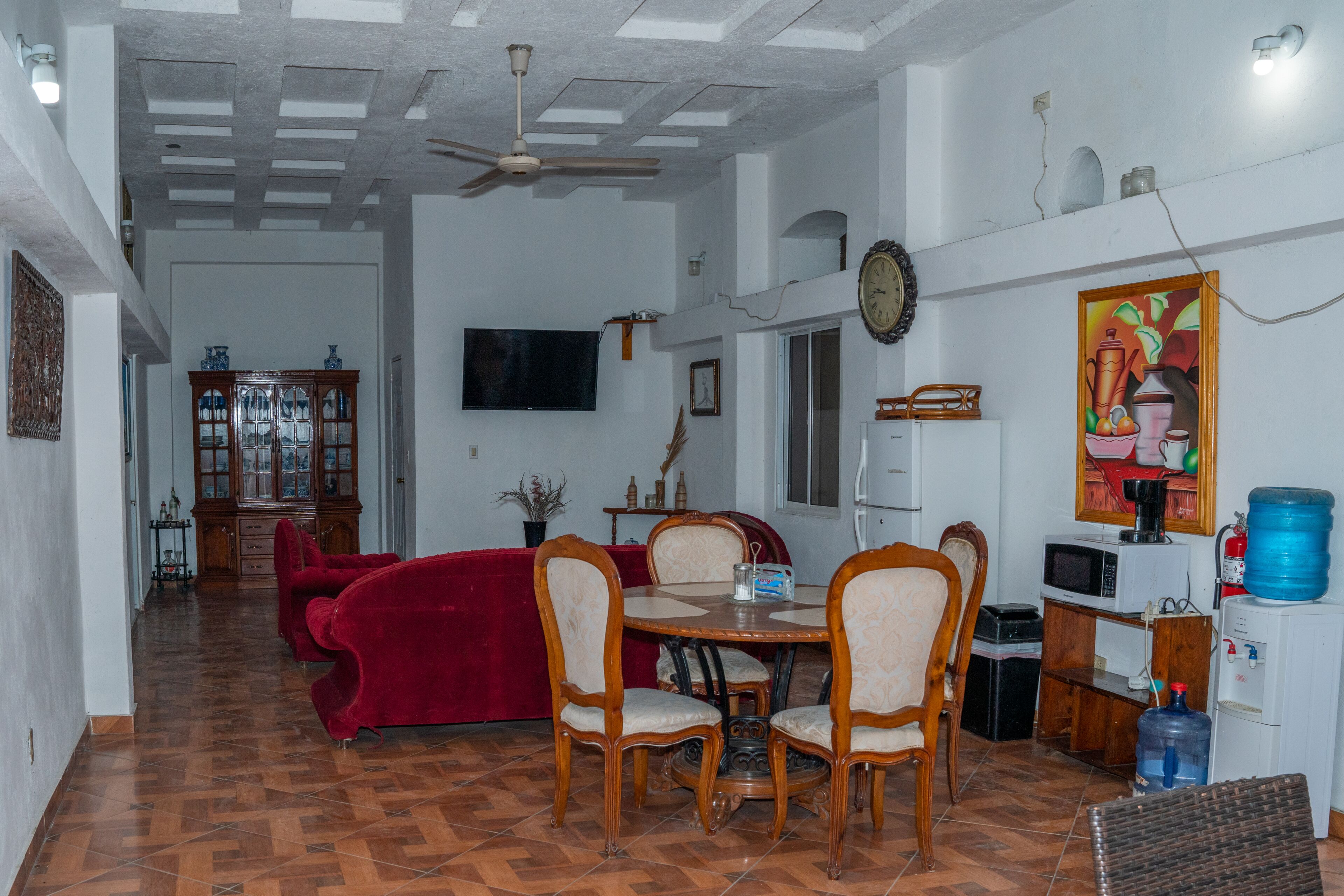 Uma sala de jantar com sofá vermelho, cadeiras de madeira, mesa de refeições, ventilador de teto, televisão e dispensador de água.