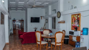 Un coin repas comprenant un canapé rouge, des chaises en bois, une table à manger, un ventilateur de plafond, un téléviseur et un distributeur d’eau.