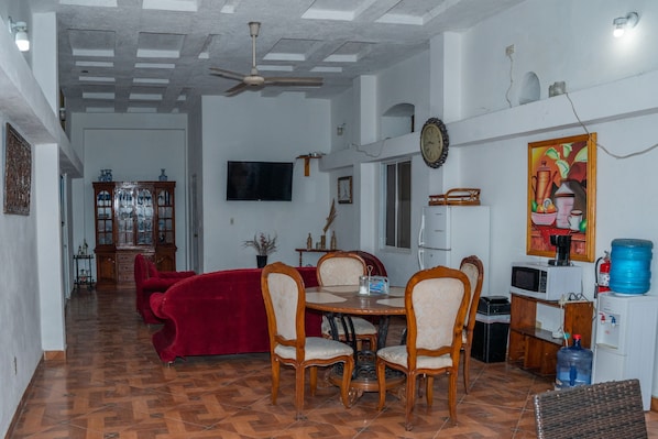 A dining area with a red sofa, wooden chairs, a dining table, a ceiling fan, a television, and a water dispenser.