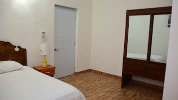 A bedroom with a wooden floor, a bed with white bedding, a wooden nightstand with a lamp, a white door, and a wooden wardrobe with a mirror.