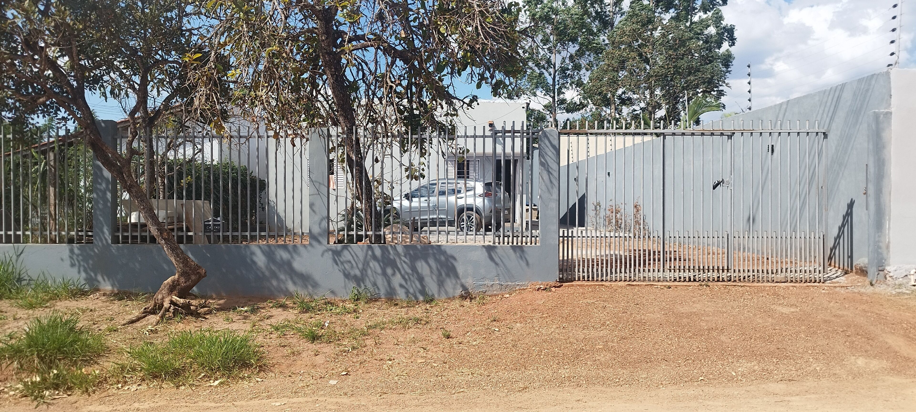 Área cercada com um carro estacionado, uma árvore e um edifício ao fundo.