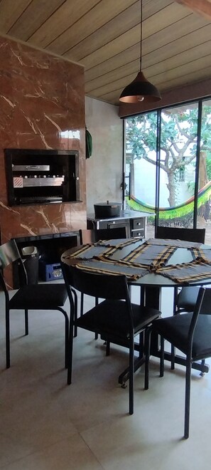 A dining area with a round table, chairs, a built-in grill, and a view of greenery outside.