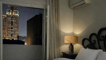 Ett hotellrum med en stor säng, ett nattduksbord, en lampa, en luftkonditioneringsanläggning och ett fönster med utsikt över stadsbilden.