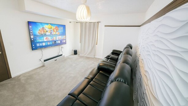 A room with a flat-screen TV mounted on the wall, a row of black leather sofas, a white textured wall, and a ceiling light fixture.