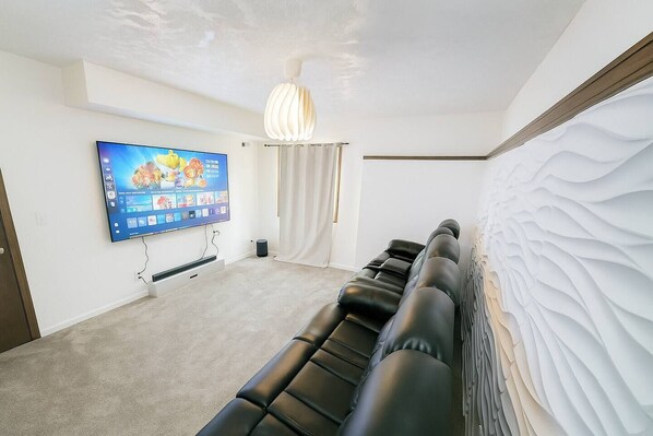 Una habitación con un televisor de pantalla plana montado en la pared, una fila de sofás de cuero negro, una pared blanca con textura y una lámpara en el techo.