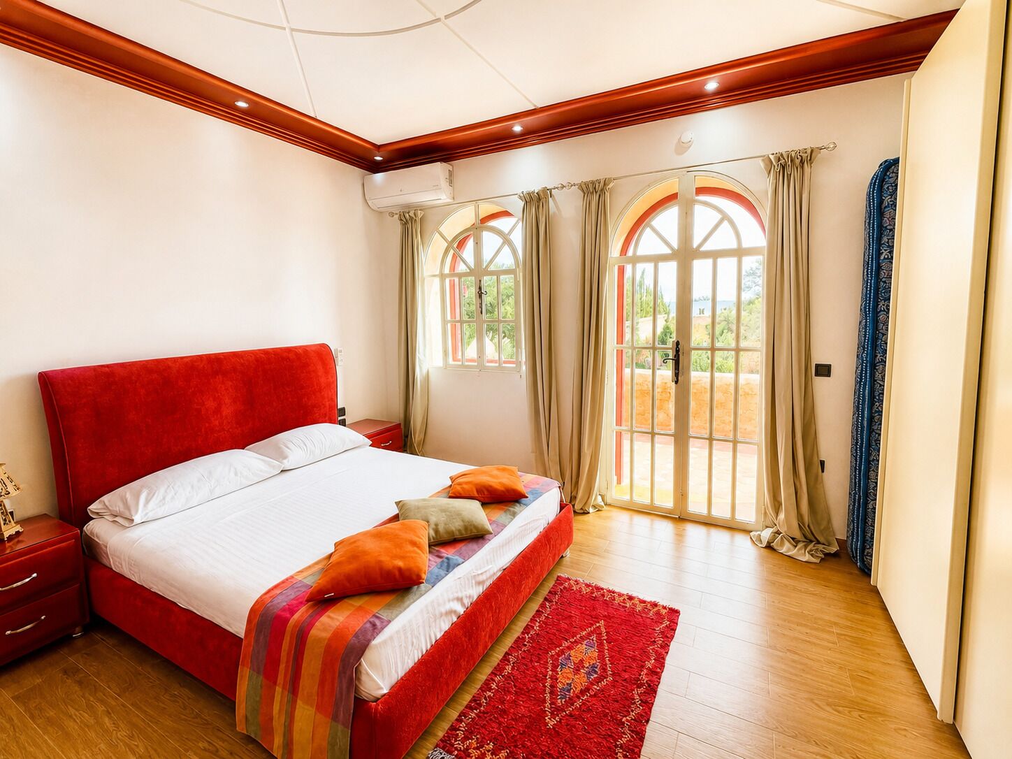 Quarto com cama vermelha, piso de madeira e vista por janelas em arco.