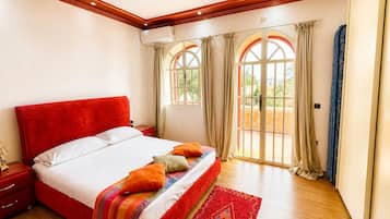 Quarto com cama vermelha, piso de madeira e vista por janelas em arco.