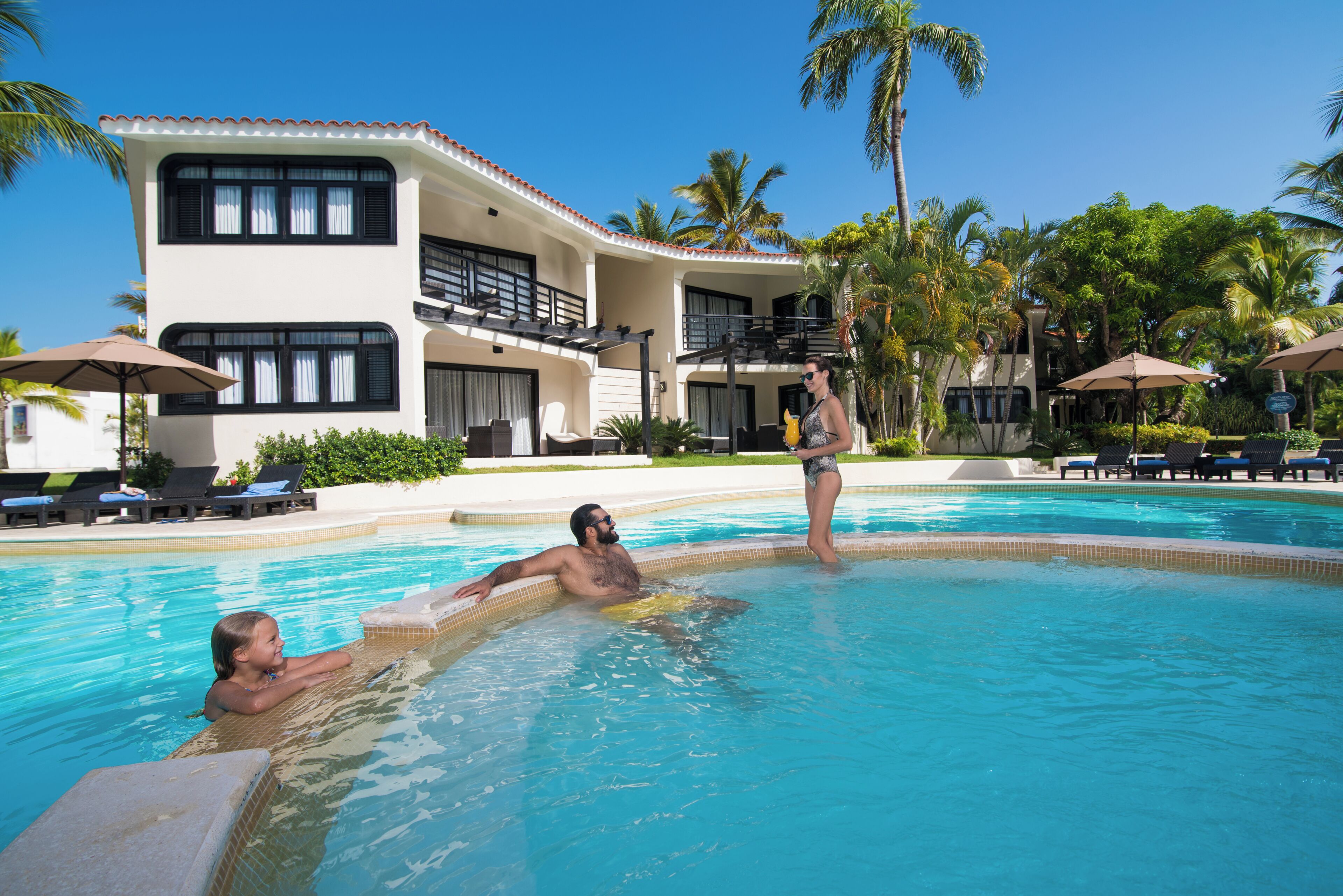Un área de piscina con sillones de descanso y sombrillas, un edificio de dos pisos con balcones, y palmeras.