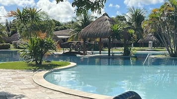 Ein Swimmingpool mit einer strohgedeckten Struktur im Hintergrund, umgeben von viel Grün und einem gefliesten Weg.