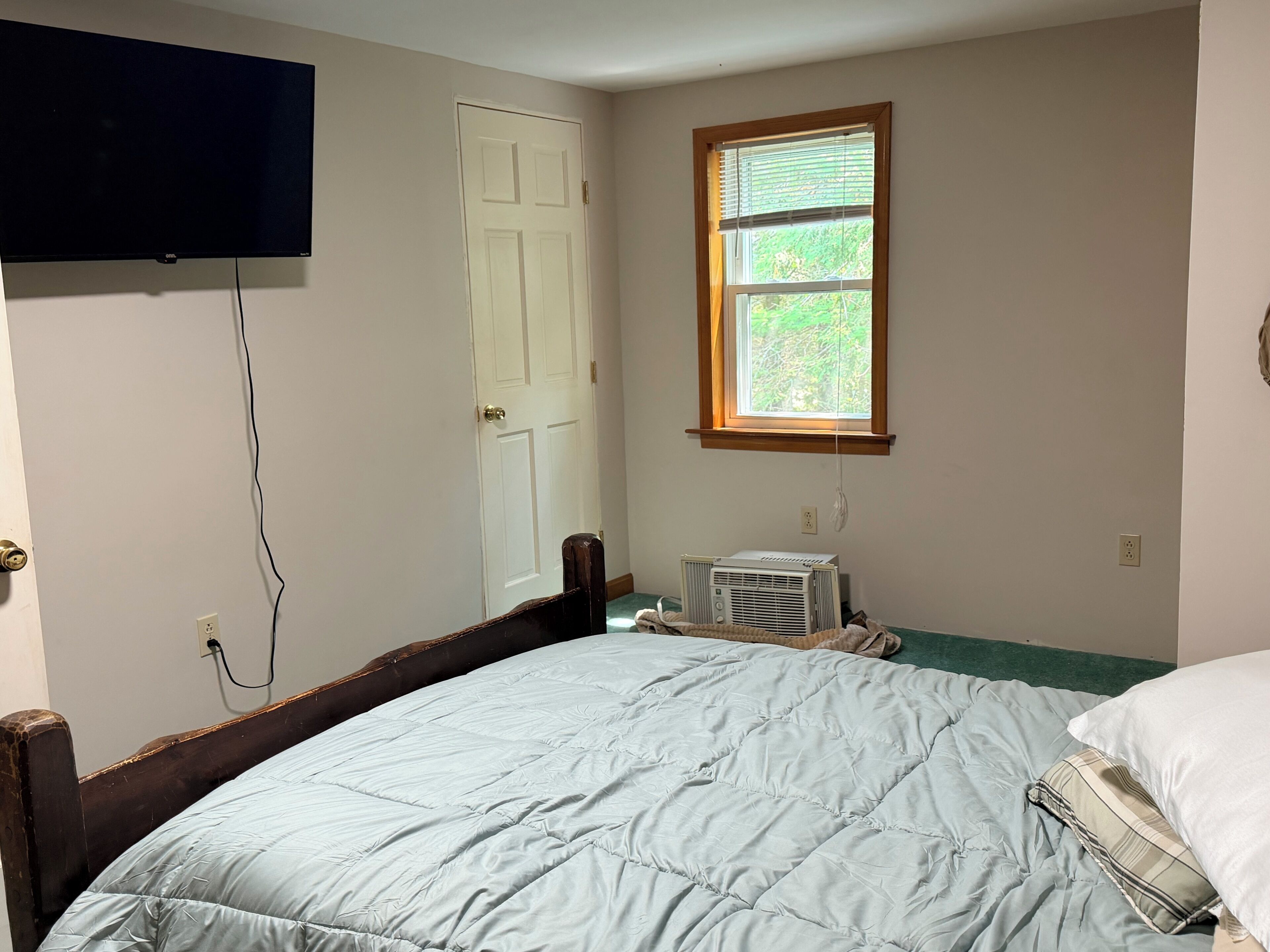 A room with a bed, a television mounted on the wall, a window with blinds, and a door.