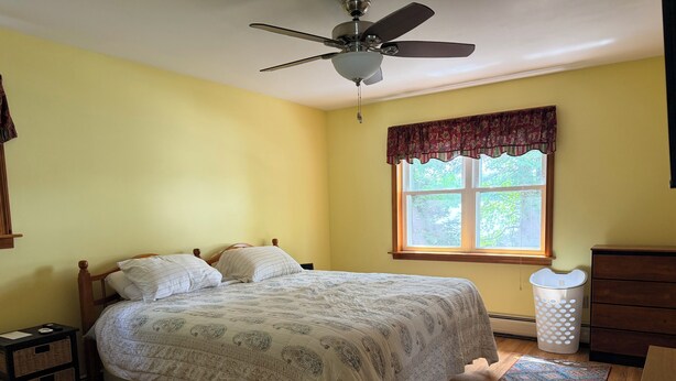Ein Schlafzimmer mit Bett, Deckenventilator, Fenster mit Vorhängen, Kommode und Mülleimer.