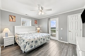 Un dormitorio con cama, mesitas de noche, ventilador de techo, ventana con persianas, puerta y vistas al exterior.