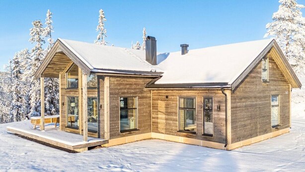 A modern wooden cabin with large windows and a covered porch, surrounded by snow-covered trees.