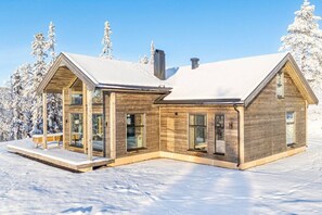 A modern wooden cabin with large windows and a covered porch, surrounded by snow-covered trees.