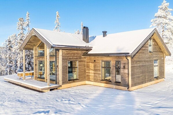 A modern wooden cabin with large windows and a covered porch, surrounded by snow-covered trees.