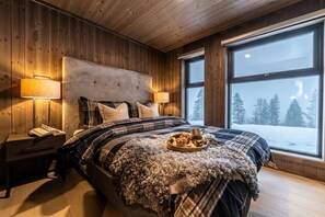 A cozy bedroom with a large bed, wooden walls, and a view of a snowy landscape through a large window.