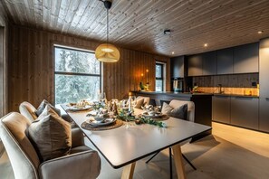 A modern dining area with a long table set for a meal, wooden walls, and a kitchen in the background.